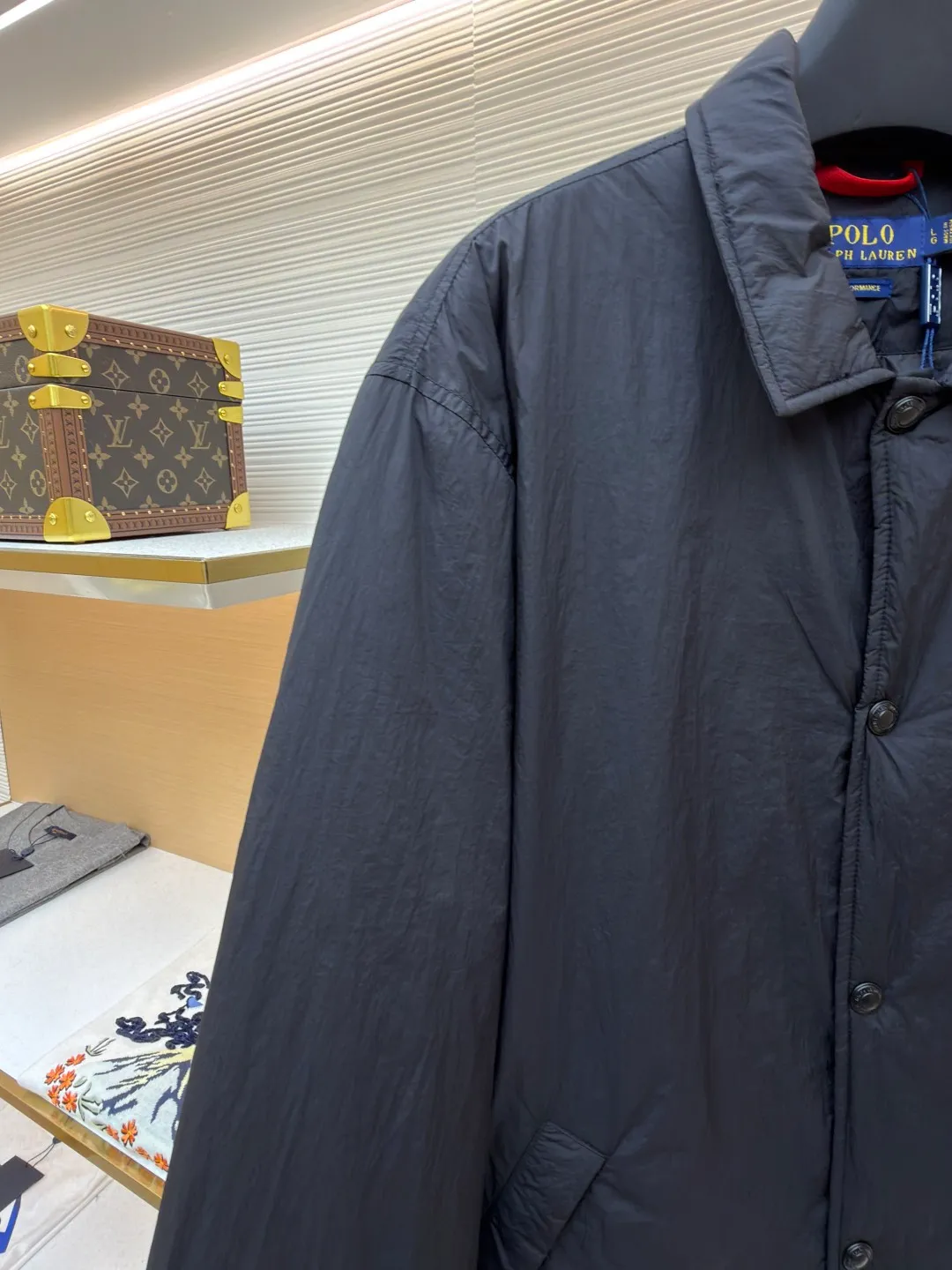 Ralph Lauren red label light cotton jacket (coach jacket) ●Original version: purchased and developed for 2699 ●Item number: 1681910-B90 ●Fabric: high-density nylon taffeta fabric ●Technology: embroidery production ●Color: black ●Version: fitted version ●Weight: about 340 grams ●Size: S-M-L-XL ●Description: The density of high-density nylon taffeta is about 210T-300T, with a smooth and slightly matte surface, excellent windproof and waterproof properties (with DWR water-repellent treatment), strong wrinkle resistance and easy care, in line with the positioning of American casual "durable and practical". Classic dark black, using vat dyeing process (dye penetrates into the fiber), the color fastness reaches ISO level 4 or above, preventing fading or color transfer after washing. The lightweight cotton-polyester blend lining (65% Polyester + 35% Cotton) reduces the friction between the fabric and inner clothing, improving the smoothness of wearing; the blended material combines breathability and durability to avoid the stuffy feeling of pure nylon lining. The classic Coach Jacket has a loose fit with slightly dropped shoulder lines (shoulder width is 1-2cm wider than the standard fit). The length reaches the hips and the sleeves are slightly tapered straight sleeves, taking into account both freedom of movement and casual style. Polo horse logo on the left chest: flat satin stitch embroidery (satin embroidery), embroidery thread: 100% polyester high color fastness embroidery thread (the color number is consistent with the brand's standard red color card), stitch density 12-15 stitches/cm, the outline of the horse logo is clear, and the details of the rider and horse are burr-free. Embroidery positioning: precise positioning through the template (12cm from the neckline and 8cm from the placket) to ensure left and right symmetry and centering. Topstitching technology: The seams of the body are made of double-needle flat seams (needle pitch 3cm/10 needles), the stitches are smooth and straight, the stitching color is dark black (the same color as the fabric), and the stitch width is 0.5cm, which enhances firmness while forming simple decorative lines. The splicing of the front placket and the body: the placket is made of double-layer folding + flat seam splicing, with a thickness of about 0.3cm to ensure that the buttons are not easily deformed when fixed; the edges of the placket are cut with a hot knife to avoid burrs. The sleeves are spliced ​​to the body: the armholes are spliced ​​with round-head keyhole machine seams + flat seams. After splicing, the armholes have a natural curvature and no sense of tightness during movement; the height of the sleeves is about 8-10cm, which is in line with the loose characteristics of casual jackets. Main button: black urea resin button, matte surface treatment, buttonholes are hand-made keyholes (6 strands of cotton thread, keyhole density 8 stitches/mm), and the edges of the eyelets are smooth and without burrs. Collar buttons: The first button on the collar is a hidden button (diameter 12mm), which prevents the collar from opening and improves the neatness of wearing.